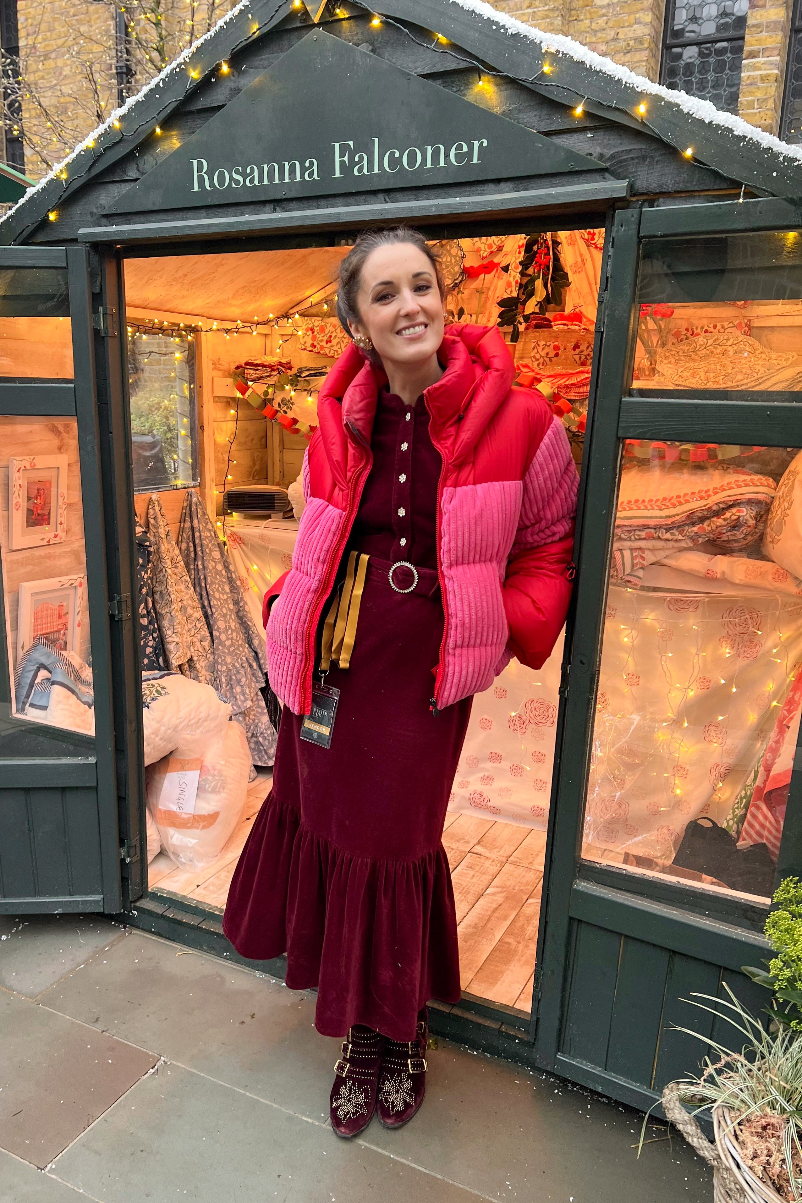 Woman in a red dress and pink puffer jacket standing inside a decorated outdoor space with 'Rosanna Falconer' branding.