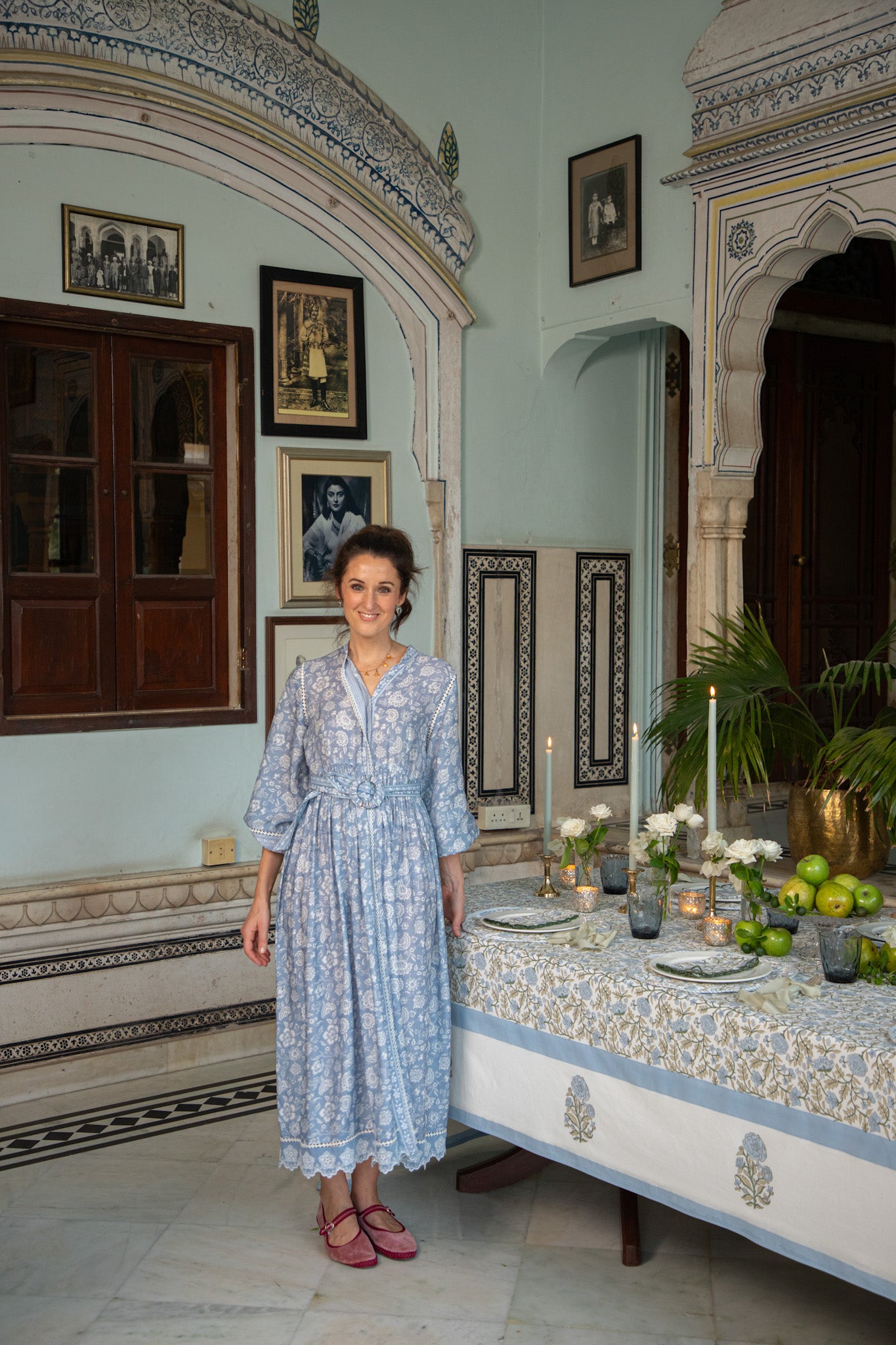 Woman in a patterned dress standing in a decorated room with a table set for a meal.