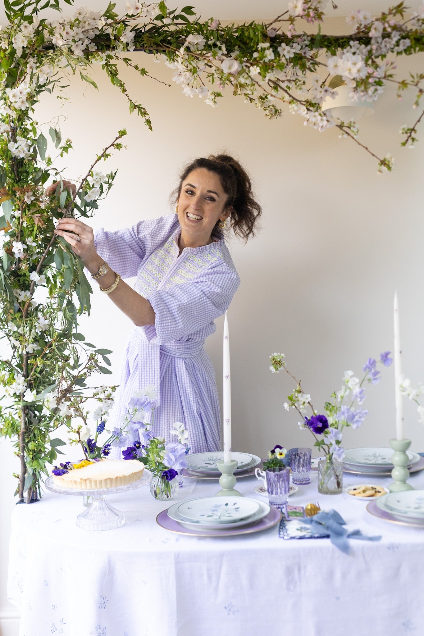 Woman in a light purple robe arranging flowers in a decorated room with floral arches and table settings.